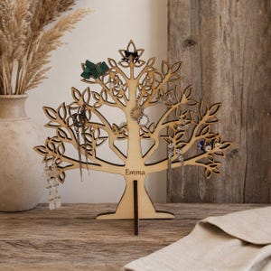 Puede incluir: Un soporte para joyas de madera clara con forma de &aacute;rbol, con ramas y hojas detalladas. La base del &aacute;rbol presenta el nombre Emma grabado. Varios pendientes y peque&ntilde;as piezas de joyer&iacute;a cuelgan de las ramas del soporte.