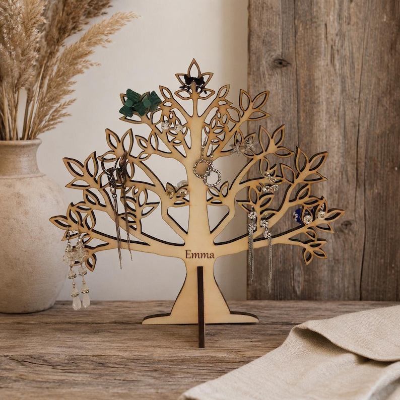 Puede incluir: Un soporte para joyas de madera clara con forma de &aacute;rbol, con ramas y hojas detalladas. La base del &aacute;rbol presenta el nombre Emma grabado. Varios pendientes y peque&ntilde;as piezas de joyer&iacute;a cuelgan de las ramas del soporte.