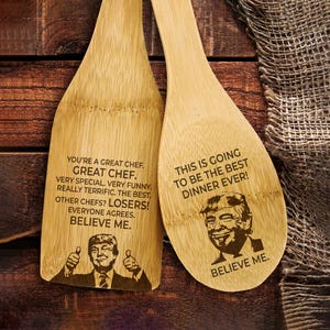 Puede incluir: Dos espátulas de cocina de madera con texto e imágenes grabados. La primera espátula dice "You're a great chef. Great chef. Very special. Very funny. Really terrific. The best. Other chefs? Losers! Everyone agrees. Believe me." y presenta una imagen de Donald Trump. La segunda espátula dice "This is going to be the best dinner ever! Believe me." y también presenta una imagen de Donald Trump.