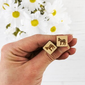 May include: Two wooden square earrings with a donkey design. The donkey is brown with white stars on its body. The earrings are held in a hand.