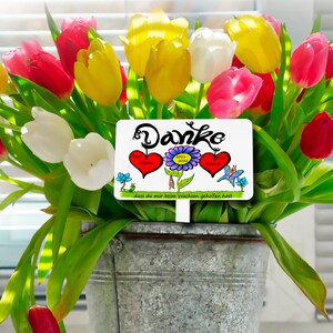 Könnte beinhalten: Ein Strauß bunter Tulpen in einem verzinkten Metalleimer. Die Blumen sind rot, gelb, rosa und weiß. Ein weißes Schild mit dem Wort "Danke" und Herz- und Blumenillustrationen befindet sich in der Mitte des Straußes.