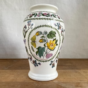 May include: A white ceramic vase with a botanical design. The vase features a detailed illustration of yellow flowers, green leaves, and a yellow butterfly, with the text "GOSSYPIUM BARBADENSE". The vase has a flared base and a rounded top.