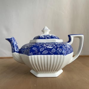 May include: A blue and white ceramic teapot featuring a floral design. The teapot has a lid and a handle. The base is white with vertical ridges. The teapot is on a wooden surface.
