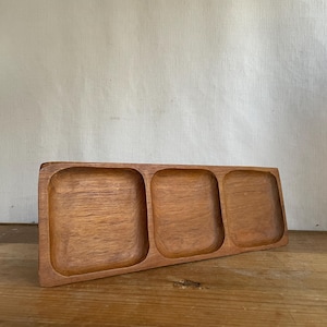 May include: A rectangular wooden serving tray featuring three distinct, square-shaped recessed compartments. The tray is crafted from a medium-toned brown wood with a visible natural grain pattern throughout the entire surface of the object.
