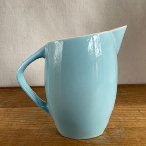 May include: A light blue ceramic jug with a smooth, glossy finish. The vessel features a rounded body, a curved handle on the left side, and a distinct, pointed pouring spout at the top rim. The design is simple, clean, and functional.