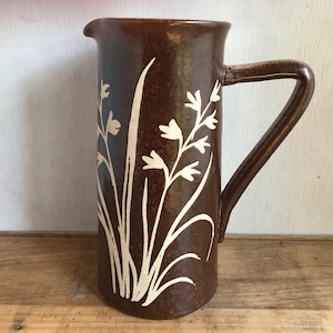 May include: A brown ceramic jug with a handle and a spout. It features a cream-coloured floral pattern. The jug is displayed on a wooden surface.
