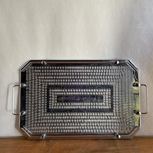 May include: A rectangular silver-toned metal serving tray featuring a polished finish and two side handles. The surface is decorated with a repeating concentric circle pattern arranged in nested rectangular borders, framed by a smooth metallic edge.