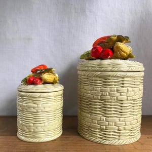 May include: Two ceramic canisters with a woven basket design. The lids are decorated with colourful fruit and vegetable sculptures, including red apples, corn, and peppers. The canisters are light green and sit on a wooden surface.