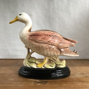 May include: A ceramic duck ornament with a white head, pinkish-brown body, and yellow beak and feet. The duck stands on a green and beige base with a black, tiered pedestal. A decorative item for the home.
