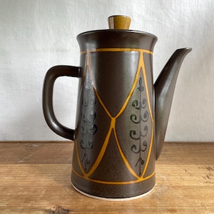May include: A brown ceramic coffee pot with a lid and handle. The pot has a geometric design with yellow and black accents. The spout is long and curved. The pot is on a wooden surface.