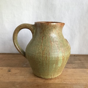 May include: A green ceramic jug with a brown rim and a handle. The jug has a textured surface and a slightly rounded shape.