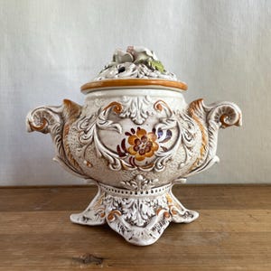 Peut inclure: Un récipient décoratif en céramique avec un couvercle, orné de motifs floraux et de volutes. Le récipient est principalement blanc cassé avec des accents orange, marron et vert. Le couvercle est surmonté d'une composition florale. Le récipient a deux poignées et un socle.