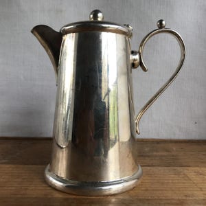 May include: A silver jug with a hinged lid and a curved handle. The jug has a polished, shiny finish.