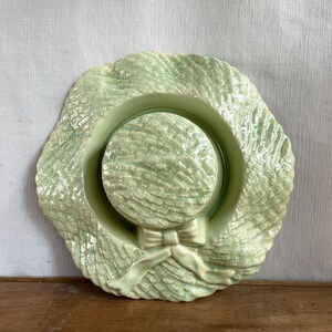 May include: A pale green ceramic hat-shaped wall decoration. The hat has a wide, textured brim and a rounded crown. A bow detail is visible on the front. This decorative item is suitable for a home or garden setting, adding a touch of vintage charm.