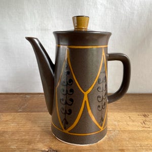 May include: A brown ceramic coffee pot with a lid and handle. The pot has a geometric design in yellow and black. The spout is long and slender, and the handle is a simple loop. The lid has a small, yellow knob.