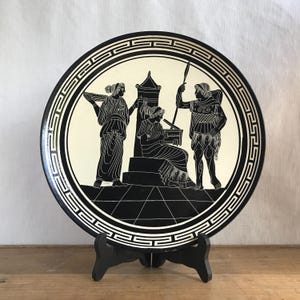 May include: Black and white ceramic plate with a Greek design featuring three figures. The plate has a white border with a repeating geometric pattern.