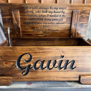 May include: A wooden toy chest with the name "Gavin" cut out on the front. The lid is open, revealing a message engraved inside. The chest is stained a warm brown color.