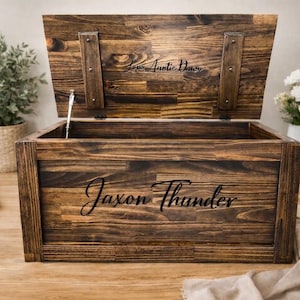May include: A wooden storage chest with a hinged lid, stained in a dark brown finish. The lid is open, revealing the interior. The name "Jaxon Thunder" is engraved on the front, and "Love Auntie Dawn" is on the lid.