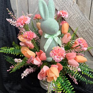 Puede incluir: Un conejito de fieltro verde pastel se encuentra en el centro de un arreglo floral. El arreglo incluye tulipanes rosas y melocot&oacute;n, claveles rosas y helechos rosas y verdes.