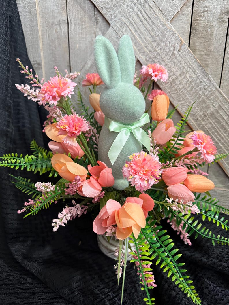 Puede incluir: Un conejito de fieltro verde pastel se encuentra en el centro de un arreglo floral. El arreglo incluye tulipanes rosas y melocot&oacute;n, claveles rosas y helechos rosas y verdes.