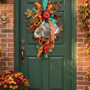 May include: A teardrop-shaped autumn wreath featuring two red pumpkins, orange and green ribbons, faux fall leaves, and two football-shaped signs. One sign reads "FAITH" and the other reads "Fall" with a football graphic and the word "FOOTBALL".