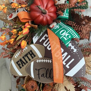 May include: A decorative autumn wreath featuring a large red pumpkin, brown football-shaped signs with the text Faith and Fall, and a large sign with the text Football. The arrangement includes orange flowers, brown leaves, and ribbons with football patterns.