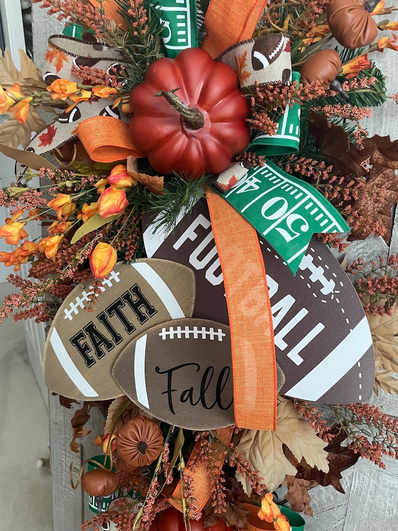 May include: A decorative autumn wreath featuring a large red pumpkin, brown football-shaped signs with the text Faith and Fall, and a large sign with the text Football. The arrangement includes orange flowers, brown leaves, and ribbons with football patterns.