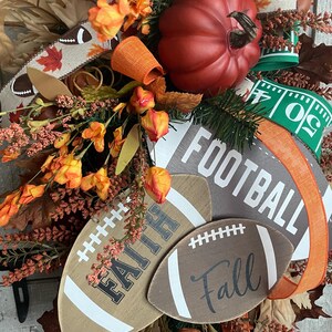 May include: A decorative wreath featuring a large red pumpkin, orange floral accents, and brown leaves. It includes wooden football-shaped cutouts with the text FAITH and Fall, along with a larger sign reading FOOTBALL, accented by ribbons.