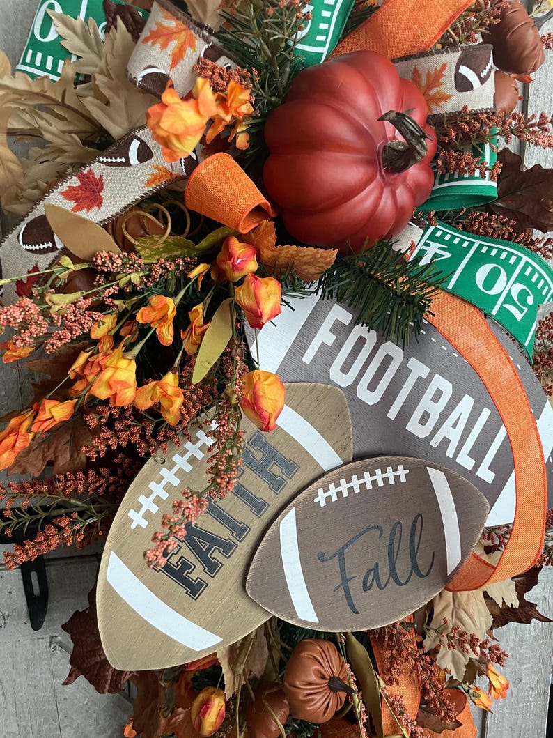 May include: A decorative wreath featuring a large red pumpkin, orange floral accents, and brown leaves. It includes wooden football-shaped cutouts with the text FAITH and Fall, along with a larger sign reading FOOTBALL, accented by ribbons.