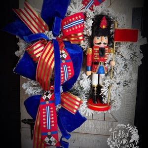 May include: A festive Christmas wreath featuring a nutcracker soldier and large blue velvet bows with red and gold striped ribbon accents. The wreath has a snowy appearance and is set against a rustic wooden door.