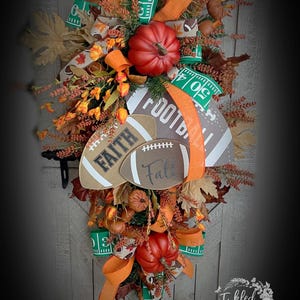 May include: A teardrop-shaped autumn wreath featuring two red pumpkins, orange ribbons, and green ribbons printed with white yard line markings. It includes three football-shaped signs with the text "FOOTBALL", "FAITH", and "Fall", accented with faux leaves.