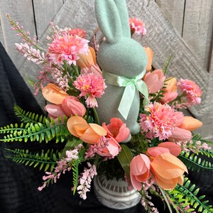 Puede incluir: Una figurita de conejo verde pastel se sienta en un jarr&oacute;n de cer&aacute;mica lleno de flores rosas y melocot&oacute;n. Las flores est&aacute;n dispuestas en un ramo primaveral.