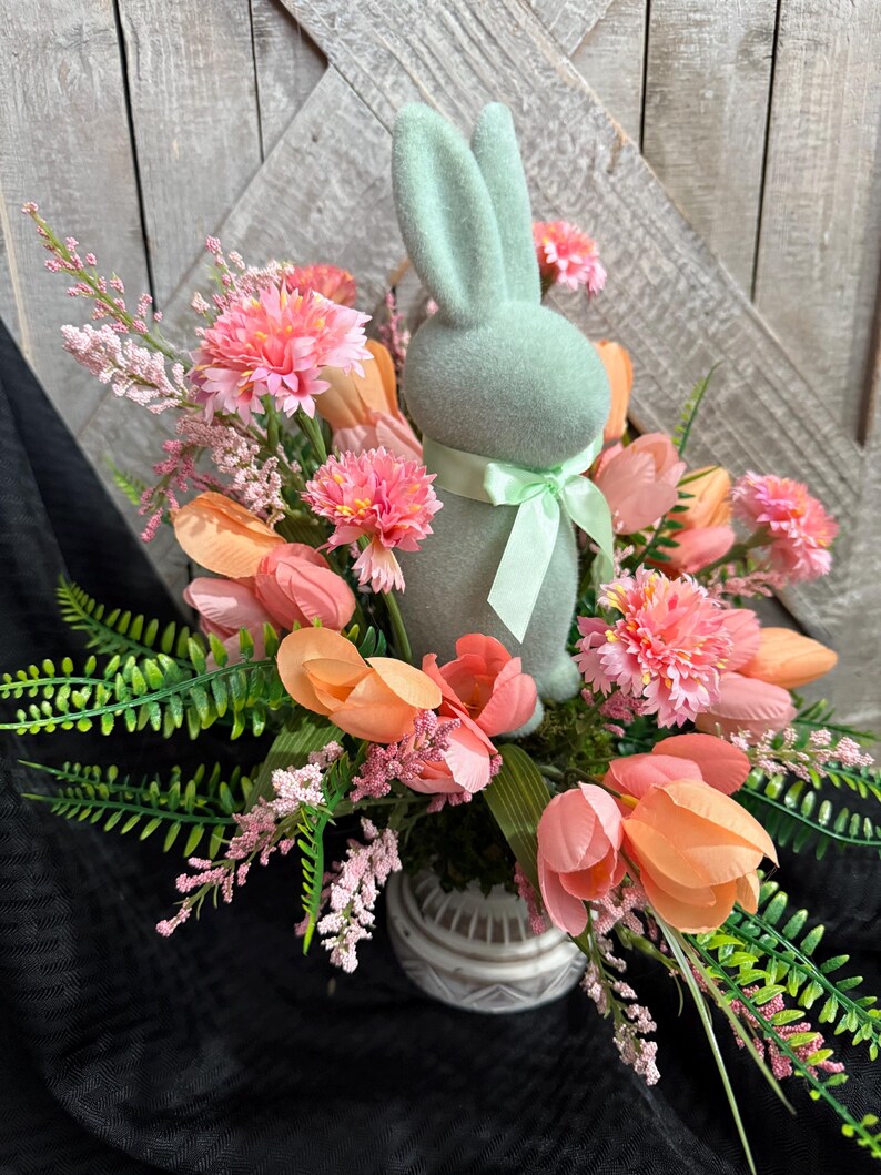 Puede incluir: Una figurita de conejo verde pastel se sienta en un jarr&oacute;n de cer&aacute;mica lleno de flores rosas y melocot&oacute;n. Las flores est&aacute;n dispuestas en un ramo primaveral.