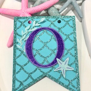 Pode incluir: Uma faixa azul turquesa com uma letra O de purpurina roxa, uma estrela do mar branca e um desenho de folhas brancas. A faixa tem um padr&atilde;o de escamas de sereia.