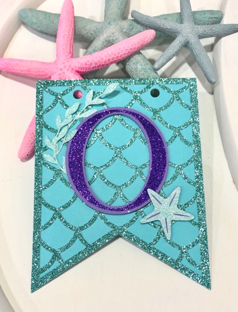 Pode incluir: Uma faixa azul turquesa com uma letra O de purpurina roxa, uma estrela do mar branca e um desenho de folhas brancas. A faixa tem um padr&atilde;o de escamas de sereia.