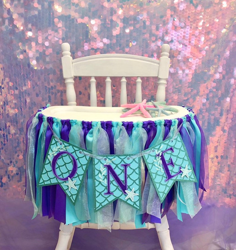 Pode incluir: Uma cadeira alta branca decorada com uma saia e uma faixa de cadeira alta com tema de sereia, em roxo, azul e verde. A faixa diz "ONE" em letras roxas com um padr&atilde;o de escamas de sereia.