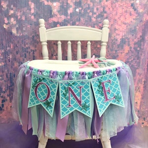 Pode incluir: Uma cadeira alta branca com uma saia de tule e fita turquesa e roxa. Uma faixa com a palavra "ONE" em letras cor-de-rosa sobre um fundo turquesa com um padr&atilde;o de escamas de sereia est&aacute; presa &agrave; saia. A faixa tem folhas verdes e detalhes de estrelas do mar.