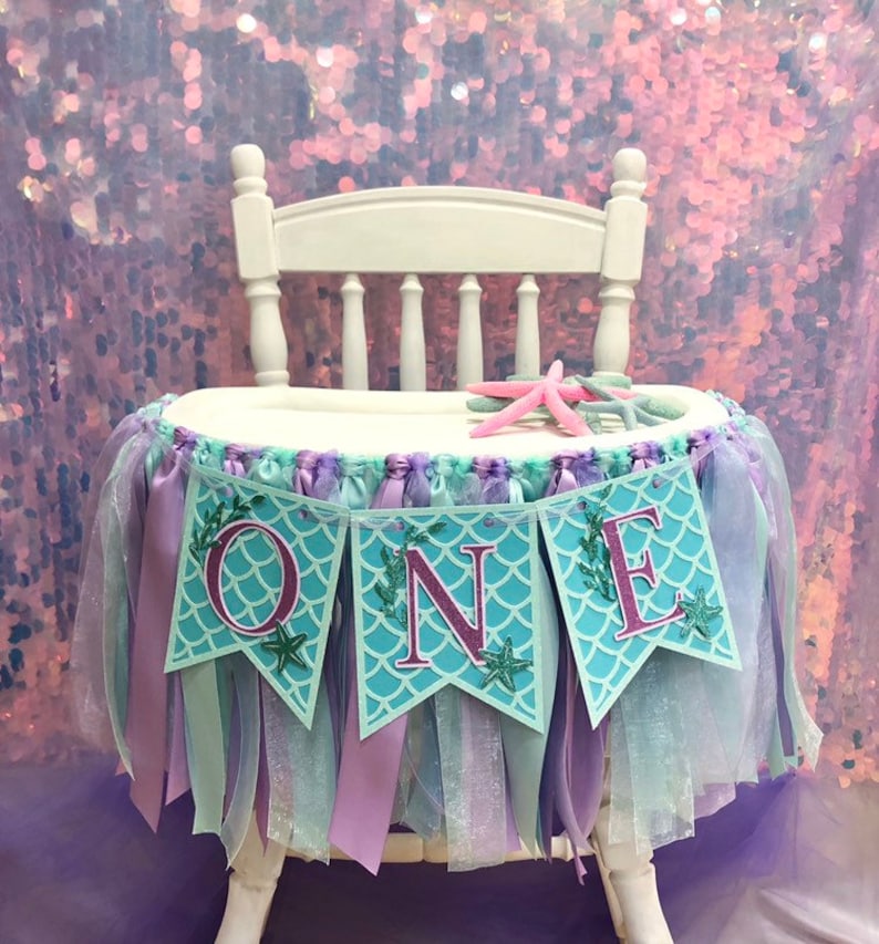 Pode incluir: Uma cadeira alta branca com uma saia de tule e fita turquesa e roxa. Uma faixa com a palavra "ONE" em letras cor-de-rosa sobre um fundo turquesa com um padr&atilde;o de escamas de sereia est&aacute; presa &agrave; saia. A faixa tem folhas verdes e detalhes de estrelas do mar.