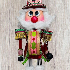 May include: A whimsical Christmas nutcracker doll with a clock-shaped hat. The doll features a pink face, white beard, and a red and green outfit. It has black and white striped legs and black boots with floral accents.
