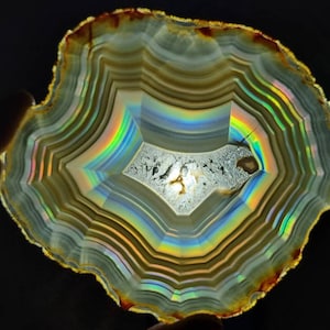 May include: A cross-section of an agate stone, showcasing concentric bands of color. The stone features a central crystalline area and a spectrum of colors, including rainbow hues, with a dark background. The stone is held by a hand.