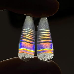 Puede incluir: Dos piedras preciosas iridiscentes en forma de lágrima con un arcoíris de colores, incluyendo azul, naranja y amarillo. Las piedras tienen una apariencia en capas y se sostienen entre los dedos sobre un fondo oscuro. La parte inferior de cada piedra tiene una textura rugosa y gris.