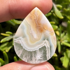 Könnte beinhalten: Ein tropfenförmiger Edelstein mit polierter Oberfläche. Der Stein weist Schichten aus Beige-, Weiß- und hellgrünen Farbtönen auf, wodurch ein Marmoreffekt entsteht. Der Edelstein wird vor einem Hintergrund aus grünem Laub gehalten.