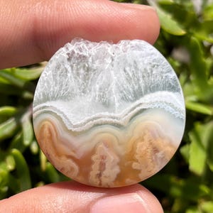Könnte beinhalten: Ein runder, polierter Edelstein mit einem natürlichen, geschichteten Muster. Der obere Teil ist weiß und kristallin, übergehend in Bänder aus Weiß, Hellgrün und Beige. Der Stein wird zwischen Fingern vor einem verschwommenen grünen Hintergrund gehalten.