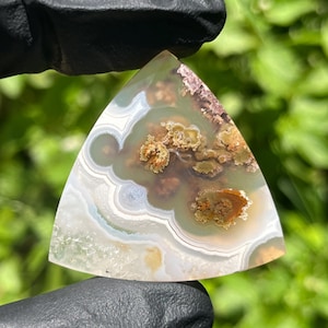 May include: A triangular-shaped polished gemstone featuring translucent light green and white banding with distinct moss-like inclusions in shades of brown, orange, and gold. The stone displays a smooth, flat surface with natural mineral patterns.