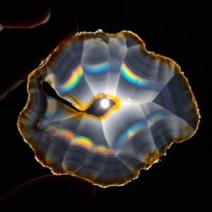 May include: A translucent, polished agate slice with a dark center and a brown edge. The stone displays a rainbow of colors from light refraction, with a dark void in the center. The agate is held against a black background.