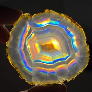 May include: A translucent, circular agate slice with a golden-brown edge. The stone displays a mesmerizing array of rainbow colours, including red, yellow, green, and blue, radiating from the centre. The agate's layered pattern is clearly visible.