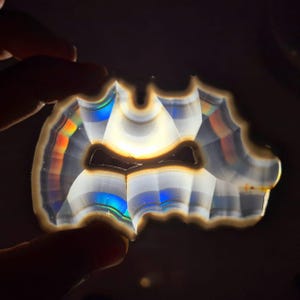 May include: A polished agate slice, held between fingers, showcasing a natural, irregular shape. The stone displays bands of white, brown, and blue, with iridescent rainbow colours. The dark background accentuates the agate's unique pattern and natural beauty.