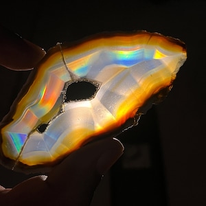 May include: A translucent agate slice with a natural, irregular shape. The stone features bands of white, orange, and brown, with iridescent rainbow colors visible throughout. A dark void is present in the center of the stone.