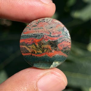 May include: A circular polished gemstone cabochon featuring a complex pattern of horizontal bands in deep forest green, vibrant orange-red, and earthy yellow tones with a smooth, reflective surface and distinct natural mineral inclusions.