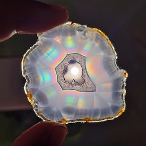 May include: A polished agate slice, translucent with a central geode. The stone shows a rainbow spectrum, with a white and grey interior and a golden-brown edge. The agate is held between two fingers.